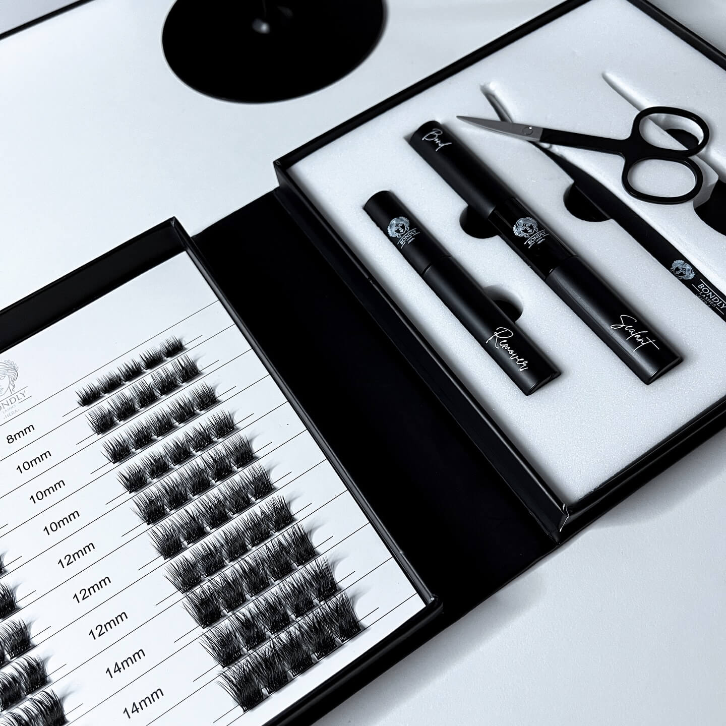 Na zdjęciu widoczny jest elegancki zestaw do aplikacji rzęs bondingowych marki Bondly Lashes, utrzymany w minimalistycznej, czarno-białej estetyce premium. W lewej części znajduje się paletka z gotowymi kępkami rzęs o różnych długościach, umożliwiająca tworzenie wielu efektów stylizacji. Po prawej stronie umieszczono niezbędne produkty do aplikacji i pielęgnacji, w tym bond, remover, sealer oraz akcesoria takie jak pęseta i nożyczki. Całość prezentuje się nowocześnie i profesjonalnie, podkreślając komfort oraz luksus domowej stylizacji rzęs.