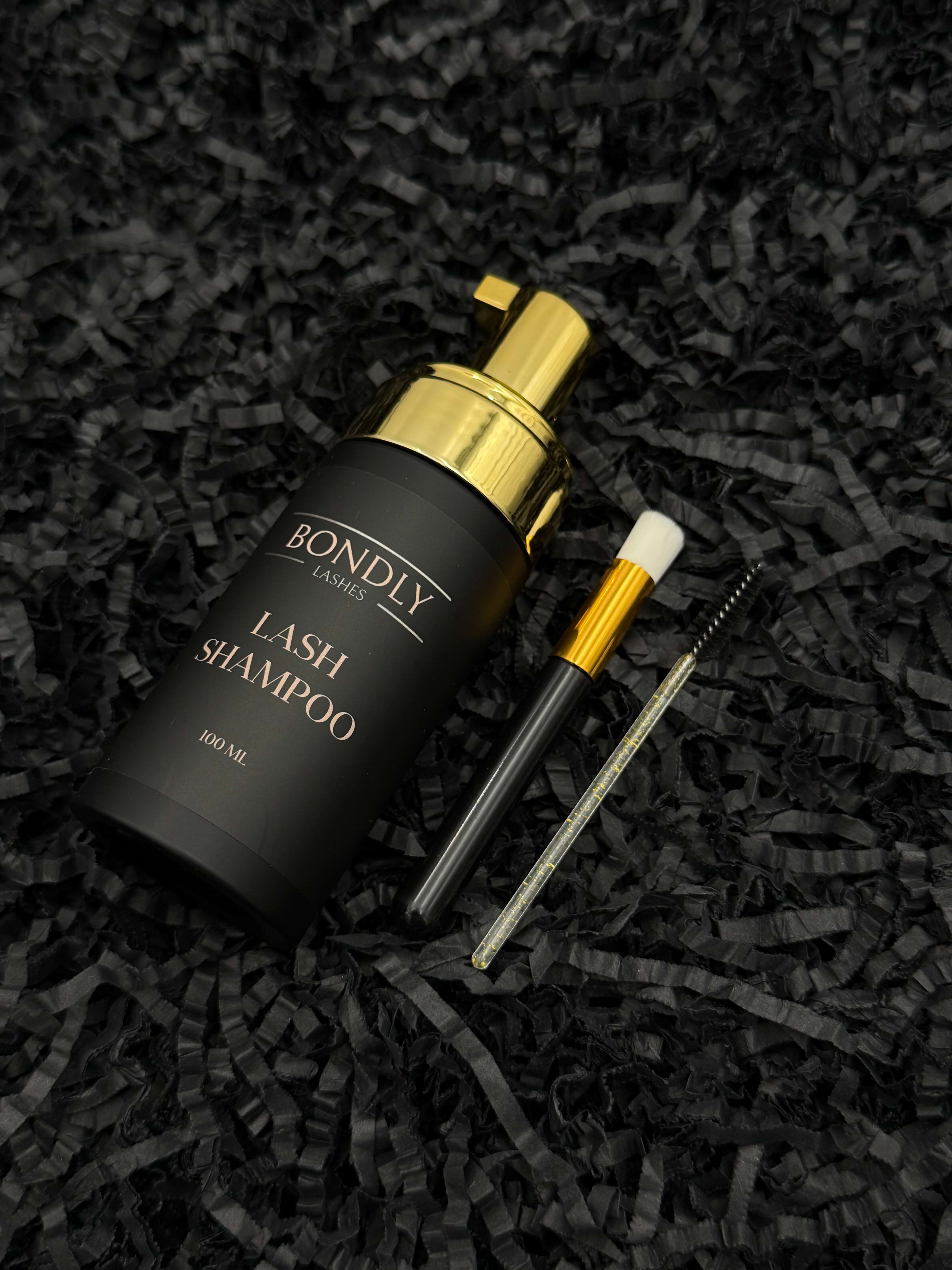 Na zdjęciu widoczny jest luksusowy szampon do rzęs marki Bondly Lashes, zaprezentowany w eleganckim czarnym opakowaniu ze złotymi detalami. Obok produktu znajdują się akcesoria do pielęgnacji i stylizacji rzęs – precyzyjny pędzelek oraz szczoteczka do rozczesywania. Całość została ułożona na dekoracyjnym, czarnym tle, które podkreśla ekskluzywny charakter produktu. Kompozycja prezentuje zestaw jako profesjonalne rozwiązanie do codziennej pielęgnacji rzęs i utrzymania ich w idealnej kondycji.